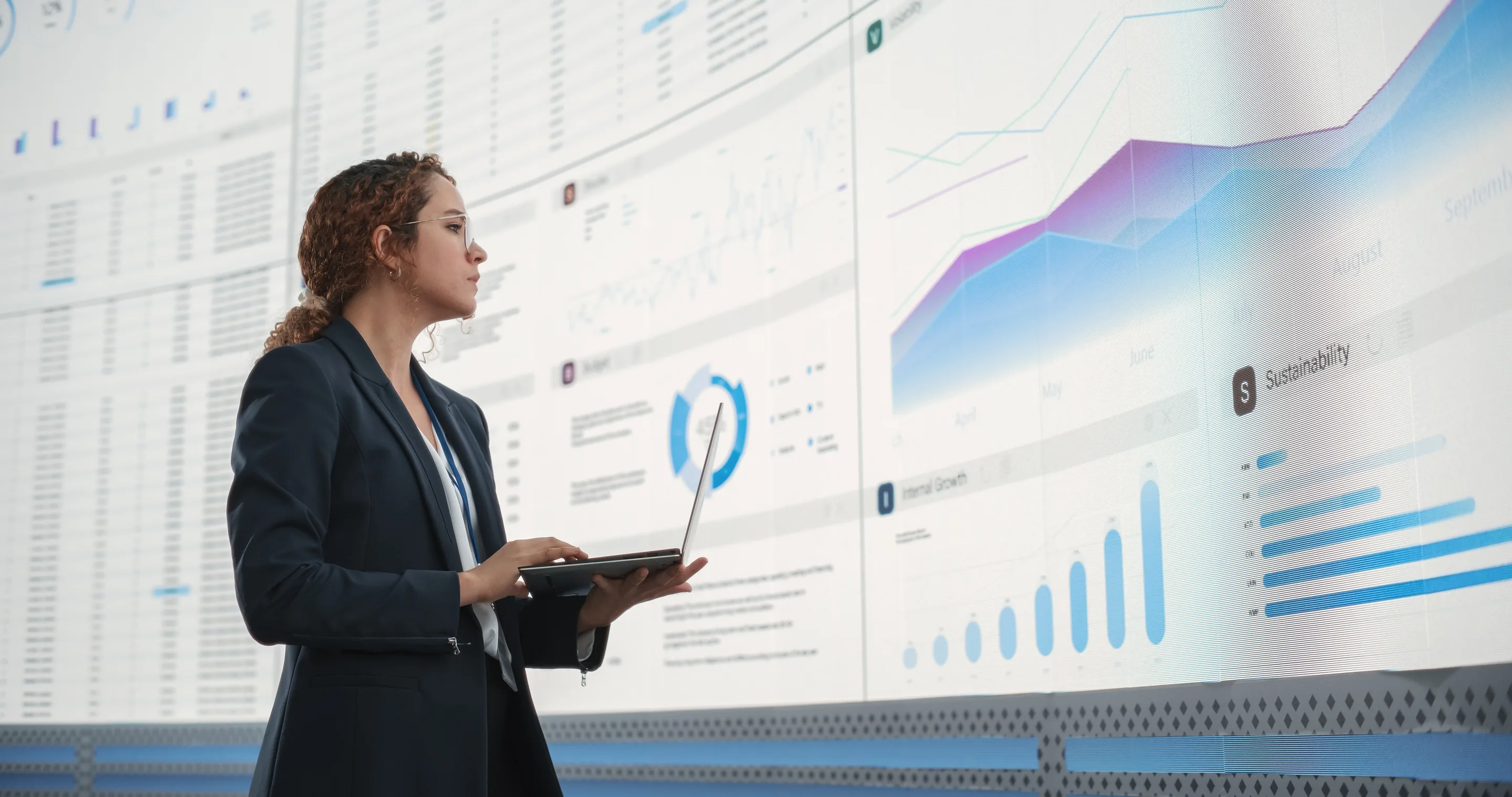 Analyst reviewing scenario results on her laptop in front of large data screens